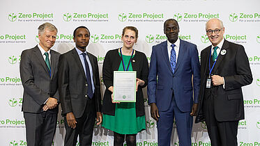 The image features five people standing in front of a backdrop with the Zero Project logo. From left to right, there is a man in a light grey suit standing next to a man in a dark suit. Next to them, a woman in a green dress and black cardigan holds a framed certificate titled "Innovative Solution 2024." She is flanked by two men, one in a blue suit and the other in a black suit, both wearing event badges. They all wear smiles and event badges.