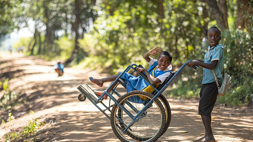 An image showing two young African children on a sunny dirt road surrounded by trees. The child in the foreground is pushing a wheelchair where the other child is seated, waving and smiling at the camera. Both are wearing school uniforms, with the child in the wheelchair in a blue dress and the one pushing in a teal shirt.