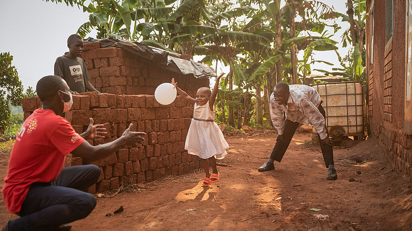 This photo shows a 3 year old girl wearing a white dress playing with a balloon with an African man wearing a CBM Tshirt.