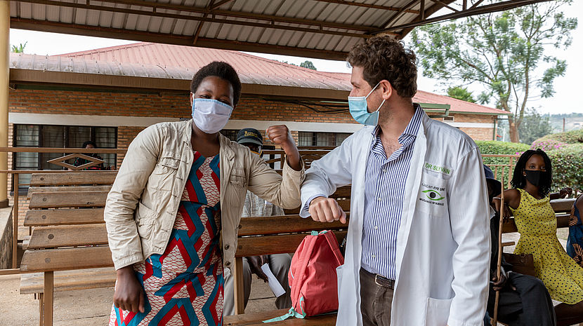 This photo shows a white doctor bumping elbows with a Black female patient wearing colourful traditional clothes.