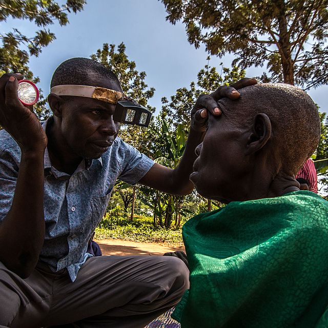 This photo shows a Ugandan man inspecting an old Ugandan woman's eyes in a field. 