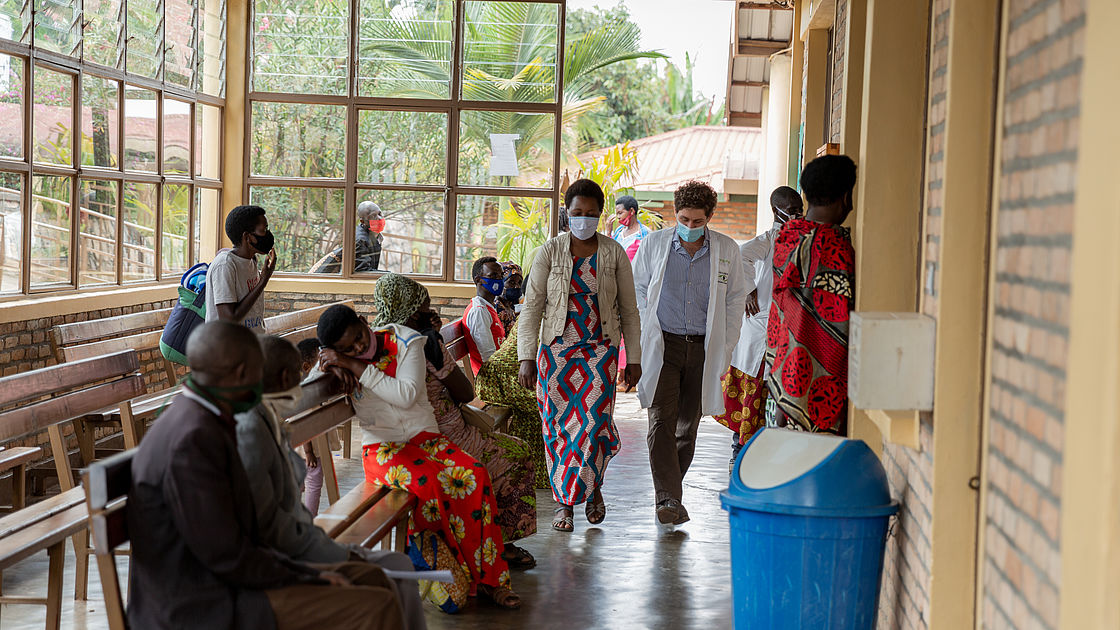 This photo shows a white doctor in conversation with his female Black patient in the corridors of the clinic, other patients are sitting on the benches nearby.