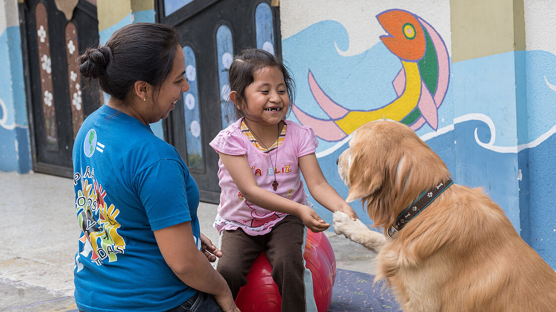 The image shows a moment of interaction in what appears to be a therapeutic or playful session with a child, a therapy dog and a facilitator. The young girl, wearing a pink T-shirt with a cheerful cartoon character, stands with the support of a large red exercise ball behind her. Facing her is a woman in a blue T-shirt, possibly a therapist, who is engaging the girl in the activity. A golden retriever therapy dog is sitting next to them, focussing its attention on the child. A wall with playful murals can be seen in the background, indicating a child-friendly environment.