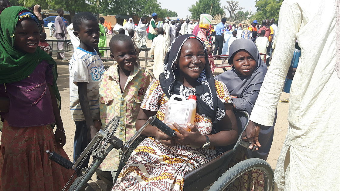 A woman smiling, holding household goods. (She uses a wheelchair)