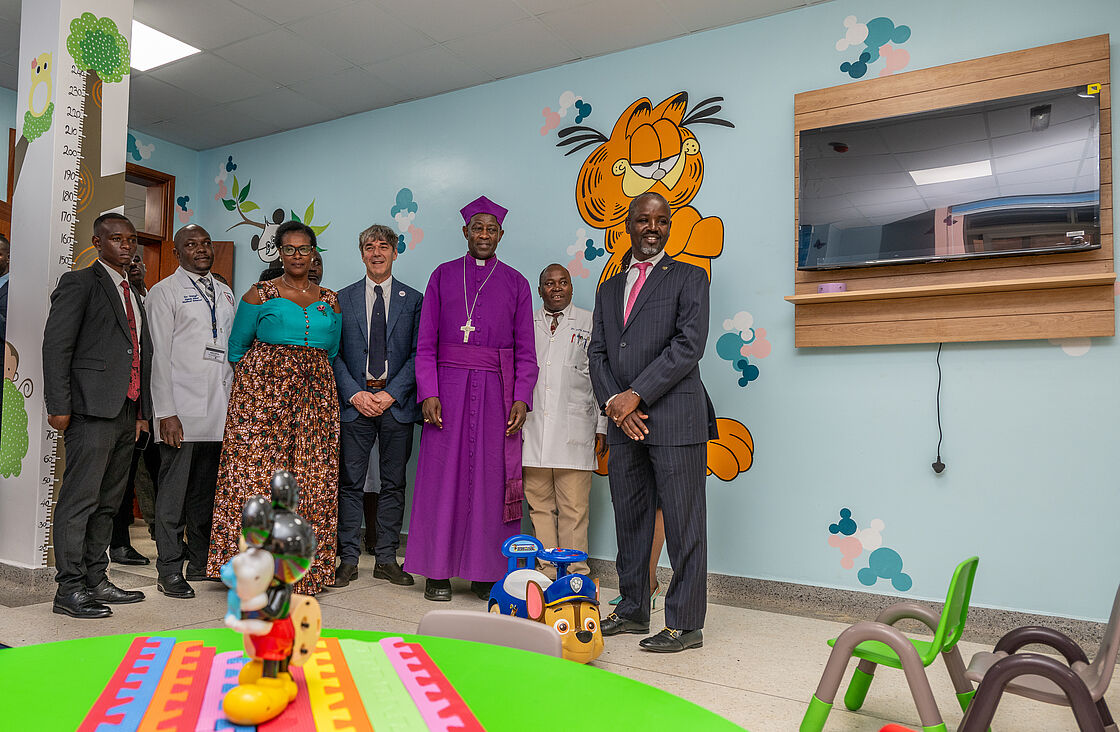The image shows a group of dignitaries and medical staff inside a brightly decorated pediatric room at the Mengo Eye Complex. The room features playful wall decorations with cartoon characters, making it child-friendly. The group includes the Archbishop of Uganda, dressed in purple clerical attire, and the Deputy Speaker of Parliament, both of whom are central figures. They are surrounded by other participants including doctors and a foreign dignitary. The atmosphere is cheerful, highlighted by the colorful furniture and educational toys in the foreground, designed to engage children during their visits. The setting demonstrates the complex's dedication to creating a welcoming environment for its young patients.