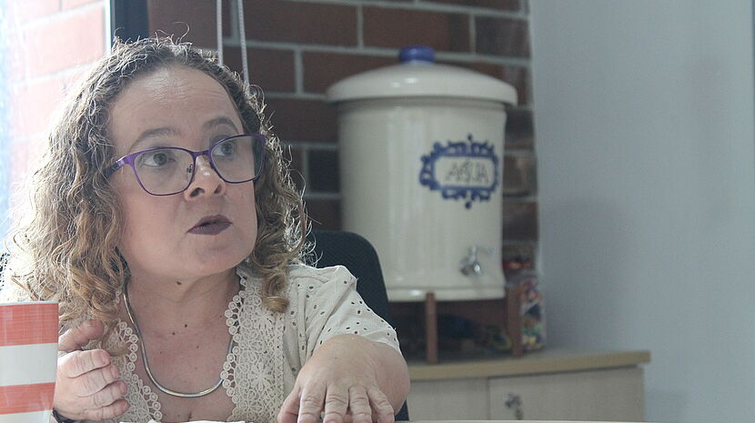 A woman with curly hair wearing glasses and a light-colored blouse with lace details is sitting at a table and speaking. She appears engaged in conversation, with her hand raised slightly for emphasis. In the background, there is a blue and white ceramic water dispenser on a wooden shelf against a brick wall, indicating a casual office setting.