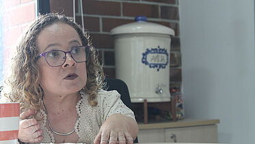 A woman with curly hair wearing glasses and a light-colored blouse with lace details is sitting at a table and speaking. She appears engaged in conversation, with her hand raised slightly for emphasis. In the background, there is a blue and white ceramic water dispenser on a wooden shelf against a brick wall, indicating a casual office setting.