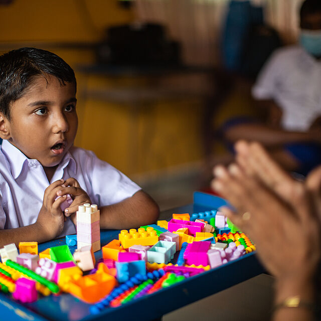 A boy playing with colourful legos in class and speaking to another person. 